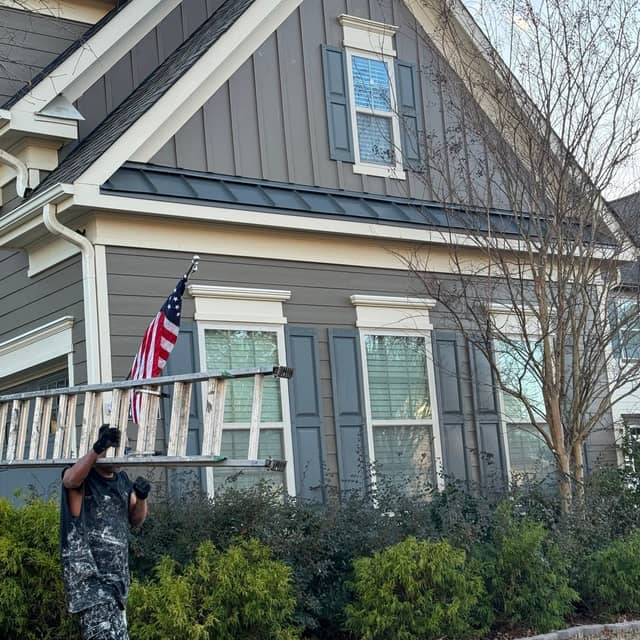 Painter carrying an extension ladder past a grey board-and-batten home with pale blue shutters